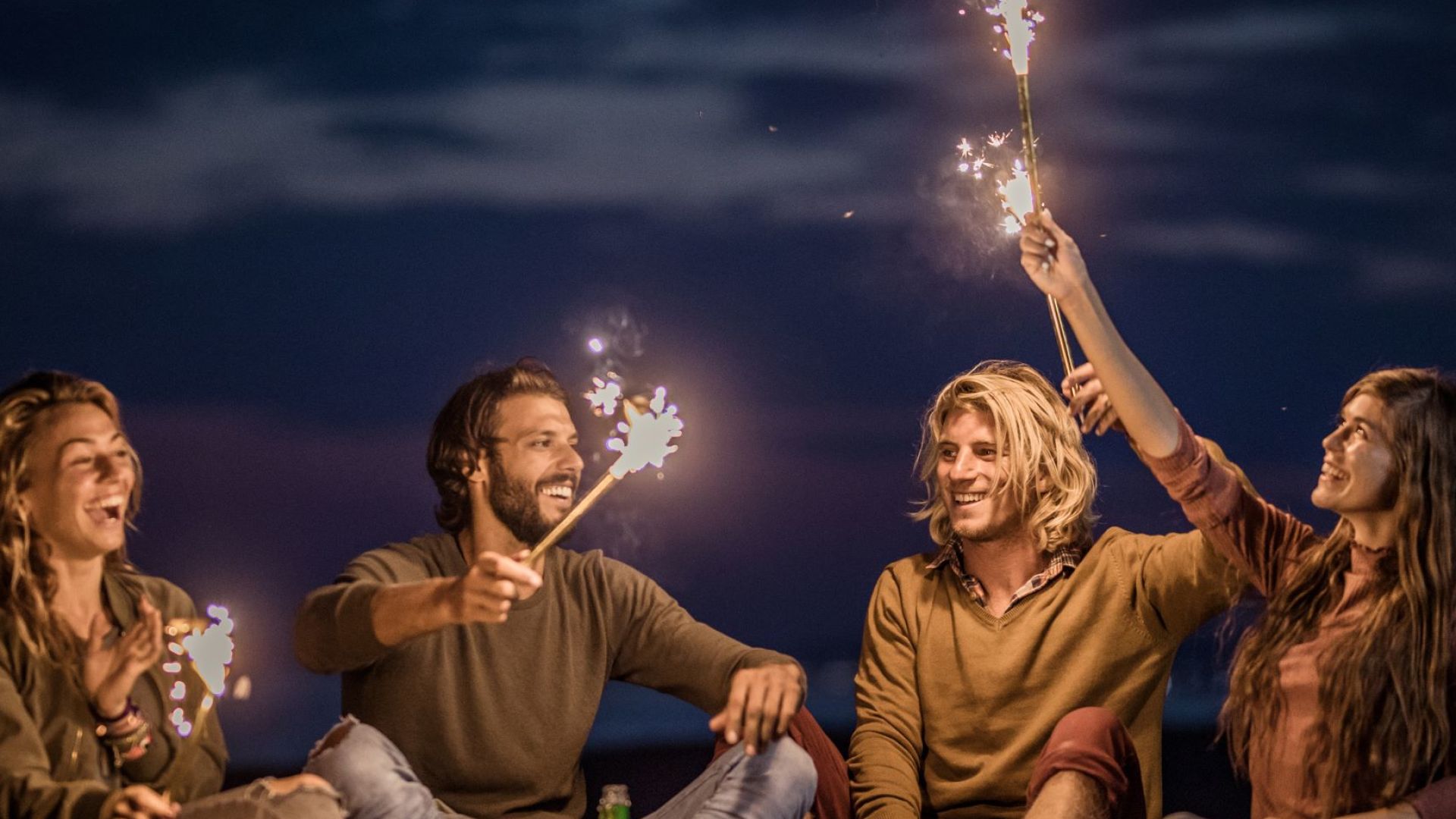 A Group Of People Sitting On A Beach With Fireworks In The Sky