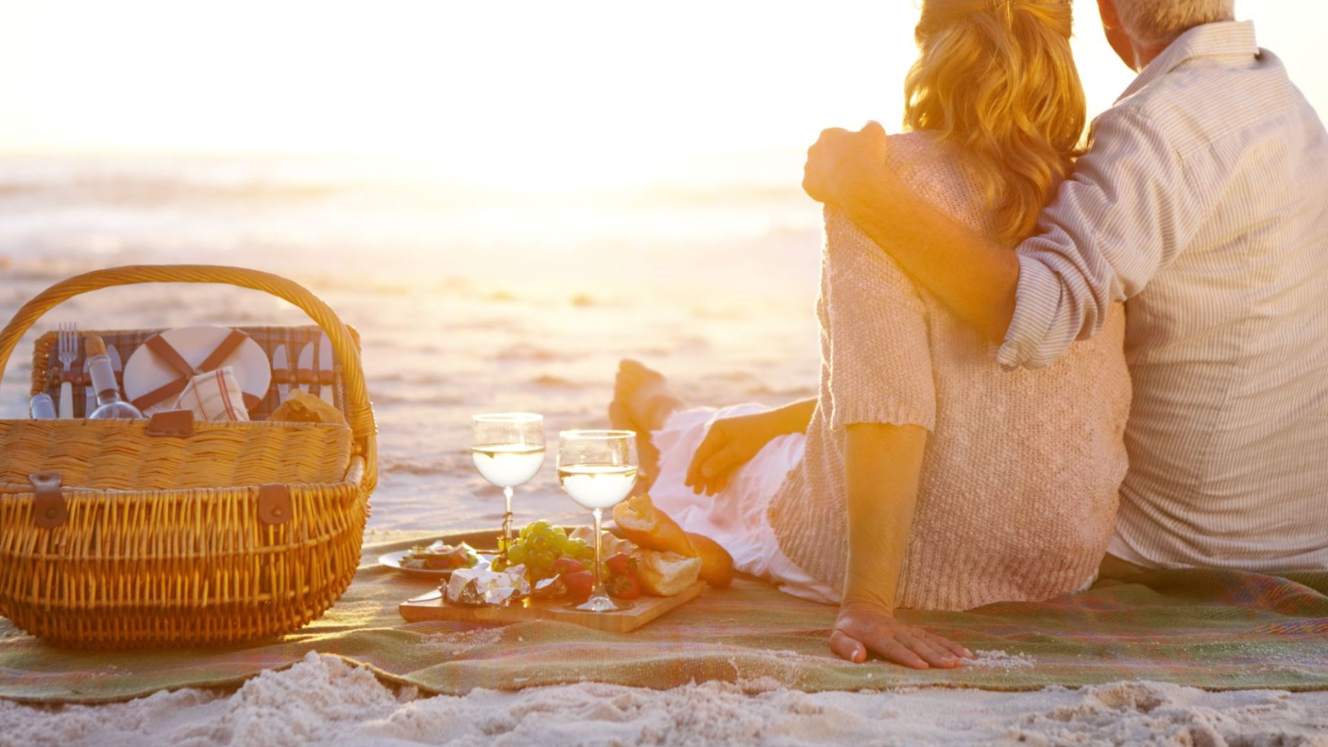 A Man And Woman Sitting On A Beach With A Basket Of Wine